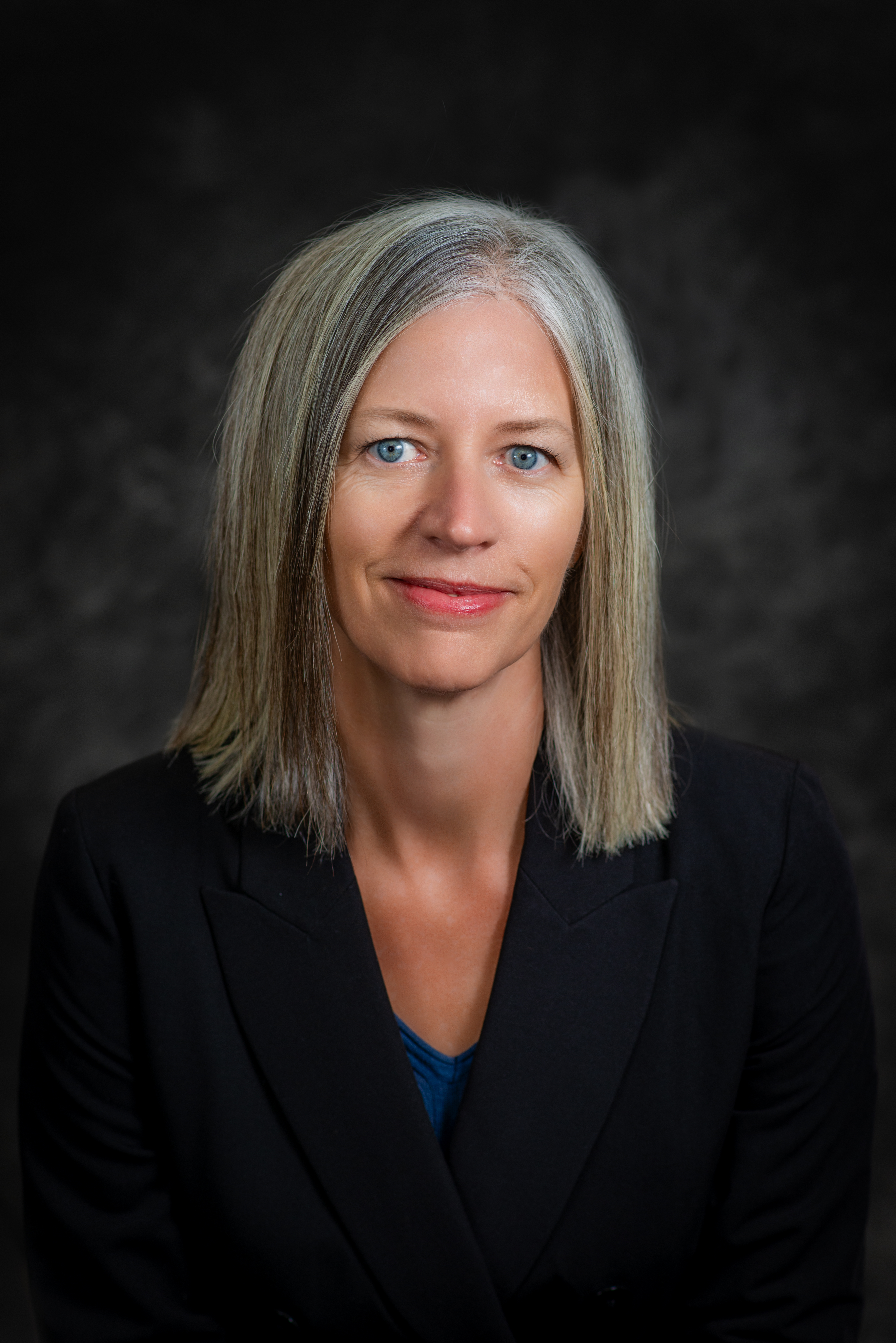 Headshot of Theresa Rowland wearing a blakc blazer with a blue shirt underneath. She has gray/blonde hair and blue eyes.