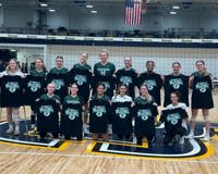 The MCC volleyball team, with players standing in the back row and players and their head coach kneeling in the front row, hold up black t-shirts while posing for a group photo.