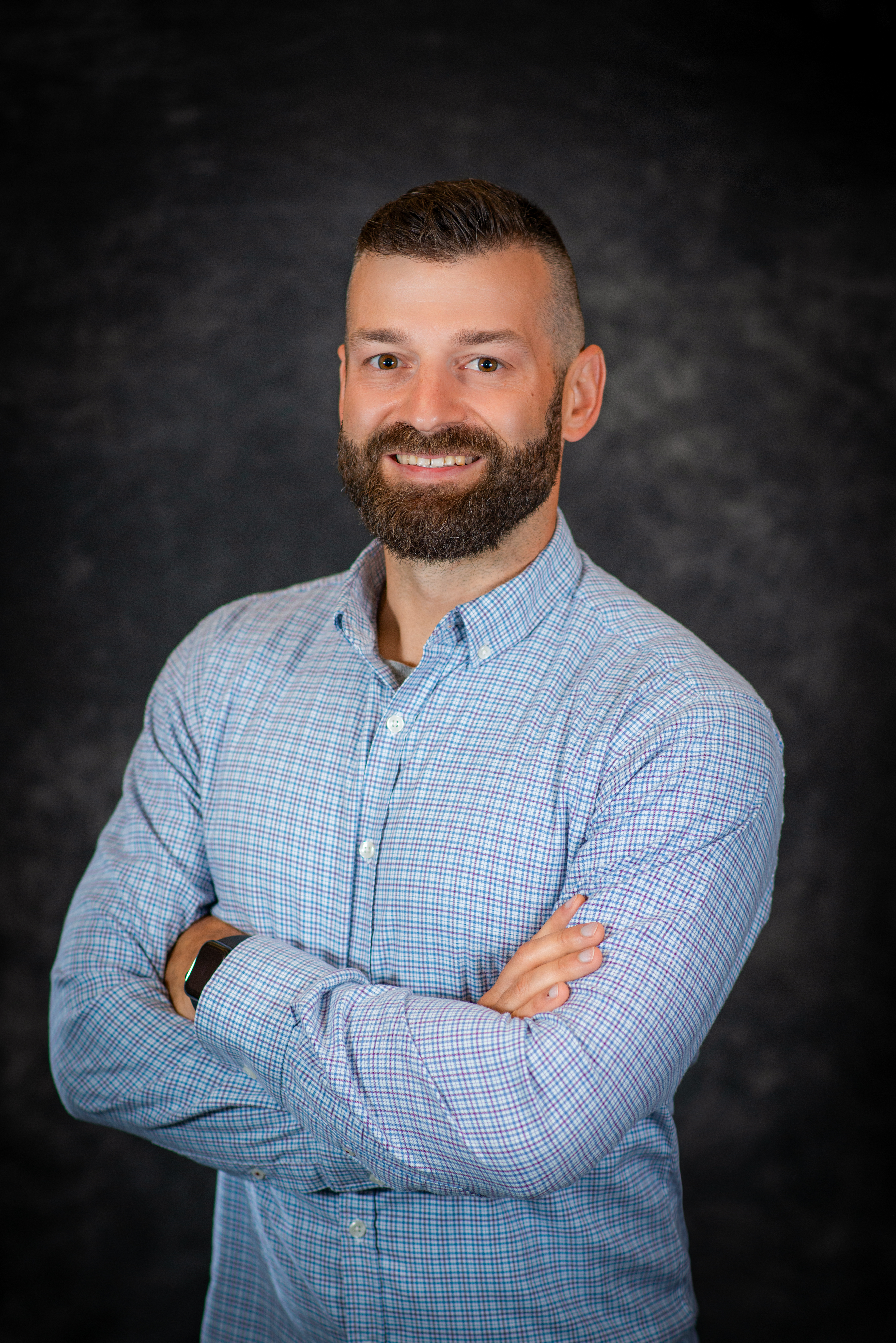 Professional photo of Taylor Male wearing a light blue plaid button down shirt in front of a gray background smiling at the camera with arms crossed.