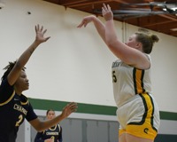 An MCC women's basketball player is photographed from the side, giving a profile view of her taking a jump shot while having it contested by a nearby defender.