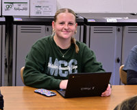 A girl wearing a green MCC sweatshirt poses for a photo while sitting at a table with her Chromebook.